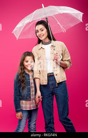 Mutter mit Regenschirm und adorable glückliche Tochter halten sich an den Händen und lächeln in die Kamera auf rosa isoliert Stockfoto