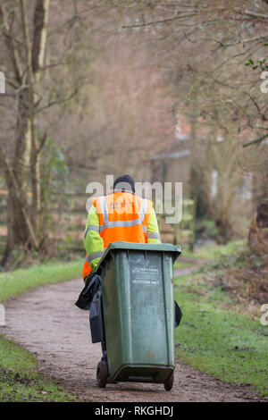 Rückansicht eines männlichen ratsarbeiters in einer gut sichtbaren Jacke, der im öffentlichen Park grüne Recycling-Mülltonnen hinter sich zieht. UK bin man, bin man bei der Arbeit. Stockfoto