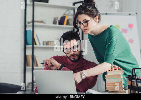 Attraktive Frau in Gläsern zeigt mit dem Finger auf Laptop in der Nähe von Menschen in modernen Büro Stockfoto