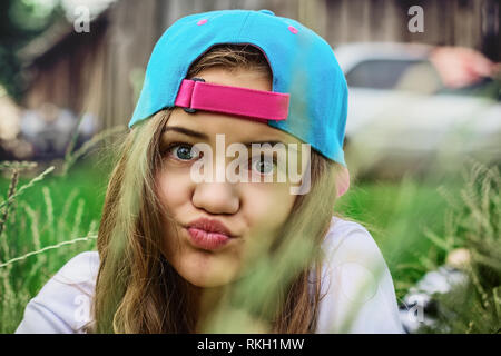 Ein jugendlicher Mädchen in einem blau Baseball cap ist, die auf dem Gras in das Dorf und ist grimassen oder Blasen einen Kuss. Ruhige Erholung im Freien. Buchse Stockfoto