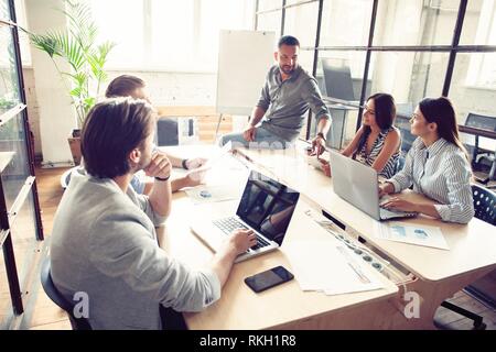 Werfen Sie einen Blick auf unsere Ergebnisse! Hübscher junger Mann stand in der Nähe der Tafel und verweist auf das Diagramm, während seine Kollegen hören. Stockfoto