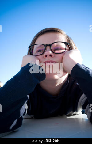 Nahaufnahme eines sitzen elf Jahre alten Jungen mit Brille sein Gesicht in seinen Händen ruht, während Tief in Gedanken und Suchen grumpy Stockfoto