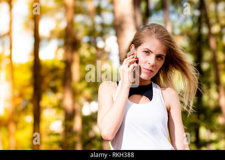 Schöne blonde Modell in der Turnhalle Kleidung Gespräch am Handy. Verschwommen Wald Hintergrund bei Sonnenuntergang Stockfoto