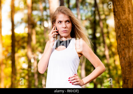 Schöne blonde Modell in der Turnhalle Kleidung Gespräch am Handy. Verschwommen Wald Hintergrund bei Sonnenuntergang Stockfoto