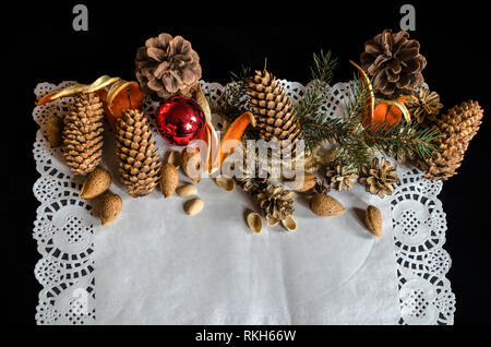 Die obere Hälfte von einem openwork Papierserviette mit verschiedenen Kegel, eine Tanne Zweig, alte abgenutzte Weihnachten Spielzeug, Mandeln und Orangen auf schwarzem Hintergrund Stockfoto