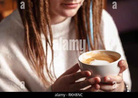 Eine weiße Tasse heißen Kaffee Latte Art in Form eines Herzens in den Händen eines jungen Mädchen mit Dreadlocks. Frau mit warmen Winter weißen Pullover gestrickt. Stockfoto