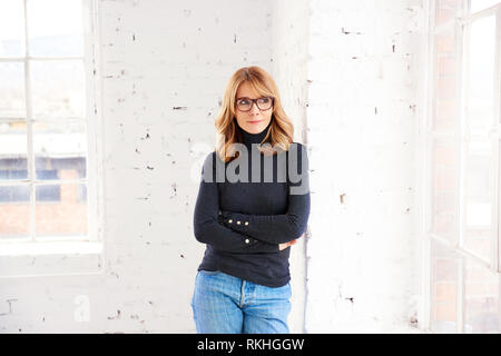 Portrait von attraktiven Frau mittleren Alters tragen Rollkragen Pullover und Jeans beim Entspannen am Fenster und Wegsehen Stockfoto
