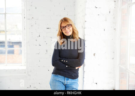 Portrait von attraktiven Frau mittleren Alters tragen Rollkragen Pullover und Jeans beim Entspannen am Fenster und Blick auf Kamera. Stockfoto