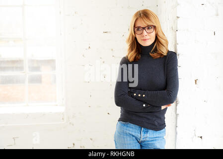 Portrait von attraktiven Frau mittleren Alters tragen Rollkragen Pullover und Jeans beim Entspannen am Fenster und Wegsehen Stockfoto