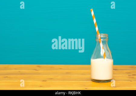Ein Drittel pint Glas Milch Flasche halb voll mit frischen Cremige Milch mit retro Papier, Stroh, auf einen hölzernen Tisch gegen eine blaugrüne Hintergrund Stockfoto