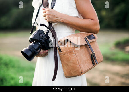 Ein Mittelteil der Frau in der Natur, die eine Kamera und eine braune Ledertasche. Stockfoto