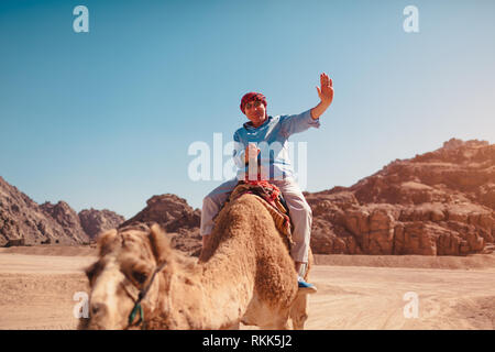 Älterer Mann reitet auf einem Kamel in der Wüste von Sinai Berge. Ihnen gerne touristische winkt für die Kamera. Urlaub in Ägypten Stockfoto