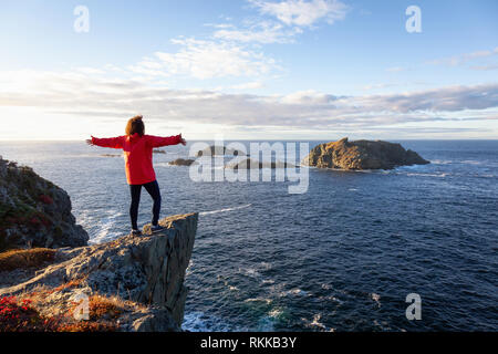 Frau in der roten Jacke steht am Rand einer Klippe mit offenen Armen und genießen das wunderschöne Meer Landschaft. Im Crow Head, North Twillingate genommen Stockfoto