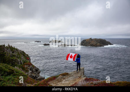 Abenteuerliche Frau mit einem kanadischen Flagge auf einem felsigen Atlantik Küste während eines bewölkten Tag. In sleepy Cove, Krähe Kopf, Twillingate, Newfoundl genommen Stockfoto