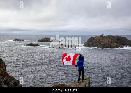 Abenteuerliche Frau mit einem kanadischen Flagge auf einem felsigen Atlantik Küste während eines bewölkten Tag. In sleepy Cove, Krähe Kopf, Twillingate, Newfoundl genommen Stockfoto