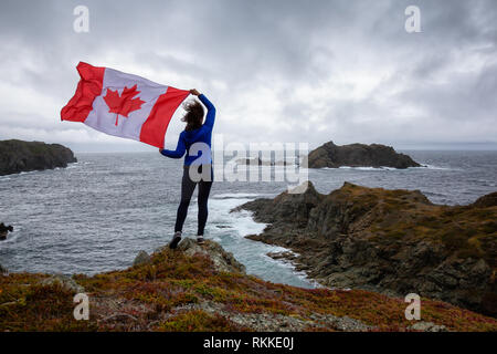 Abenteuerliche Frau mit einem kanadischen Flagge auf einem felsigen Atlantik Küste während eines bewölkten Tag. In sleepy Cove, Krähe Kopf, Twillingate, Newfoundl genommen Stockfoto