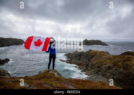Abenteuerliche Frau mit einem kanadischen Flagge auf einem felsigen Atlantik Küste während eines bewölkten Tag. In sleepy Cove, Krähe Kopf, Twillingate, Newfoundl genommen Stockfoto