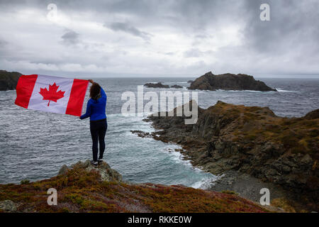 Abenteuerliche Frau mit einem kanadischen Flagge auf einem felsigen Atlantik Küste während eines bewölkten Tag. In sleepy Cove, Krähe Kopf, Twillingate, Newfoundl genommen Stockfoto