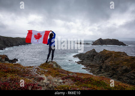 Abenteuerliche Frau mit einem kanadischen Flagge auf einem felsigen Atlantik Küste während eines bewölkten Tag. In sleepy Cove, Krähe Kopf, Twillingate, Newfoundl genommen Stockfoto