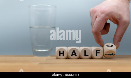 Hand einen Würfel und Änderungen der Ausdruck "halb leer" zu "halb voll". Ein halbes Glas Wasser steht vor einem grauen Hintergrund. Stockfoto