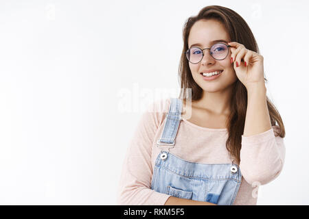 Nahaufnahme der charmante assertive Smart weibliche in Overalls die Hand auf Gläsern und lächelnd im Großen und Ganzen zufrieden mit der Kamera erreicht Stockfoto