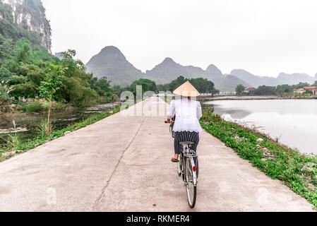 Vietnamesische Frau mit traditionellen konischen Strohhut auf dem Fahrrad. Die schöne Landschaft der Reisfelder und Bergwelt im Naturschutzgebiet Trang Ein und Stockfoto