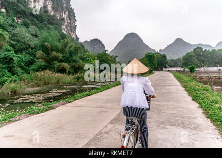 Vietnamesische Frau mit traditionellen konischen Strohhut auf dem Fahrrad. Die schöne Landschaft der Reisfelder und Bergwelt im Naturschutzgebiet Trang Ein und Stockfoto