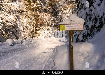 Schneebedeckten Holz- Schild mit Warnung Text in Deutsch und Englisch: Mountainbiker Sie bitte langsamer. Sonnigen Tag, Wanderweg im Winter Landschaft. Stockfoto