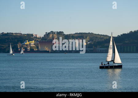 Lissabon, Portugal - 12/28/18: Segelboote auf den Fluss Tagus und Sonne auf weißem Segel, mit ruhigem Wasser, mit grossen Fabrik gebäude im Hintergrund Riverside Stockfoto