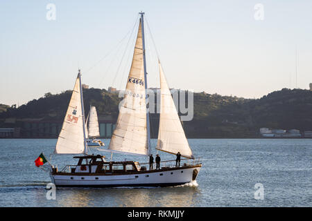 Lissabon, Portugal - 12/28/18: Segelboote auf den Fluss Tagus und Sonne auf weißem Segel, mit ruhigem Wasser, mit grossen Fabrik gebäude im Hintergrund Riverside Stockfoto