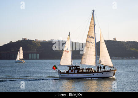 Lissabon, Portugal - 12/28/18: Segelboote auf den Fluss Tagus und Sonne auf weißem Segel, mit ruhigem Wasser, mit grossen Fabrik gebäude im Hintergrund Riverside Stockfoto