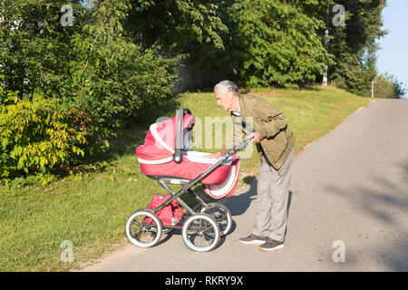 Großvater trägt Enkelin in Baby carriager bei Sonnenuntergang. Großvater lehnte sich hinüber zum Baby. Outdoor im Land. Stockfoto