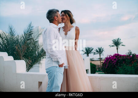 Glückliche Brautpaar Kuss auf der Terrasse des weißen Villa gegen den Hintergrund der Palmen, Zweige und Blüten während der Flitterwochen in Ägypten. Stockfoto