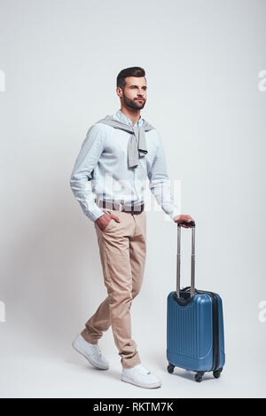 Travel Concept. Volle Länge des charmanten bärtiger Mann mit einem Gepäck beim Stehen auf einem grauen Hintergrund. Studio shot Stockfoto