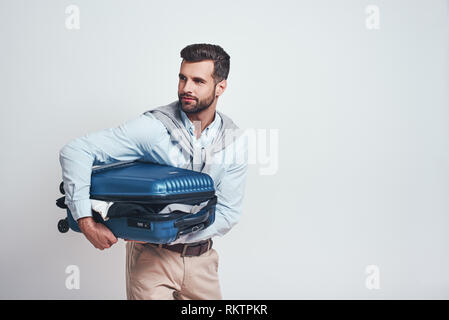 Bereit zu Reisen. Lächelnd schöner Mann hält Koffer voller Kleider beim Stehen auf einem grauen Hintergrund. Travel Concept. Close-up Portrait Stockfoto