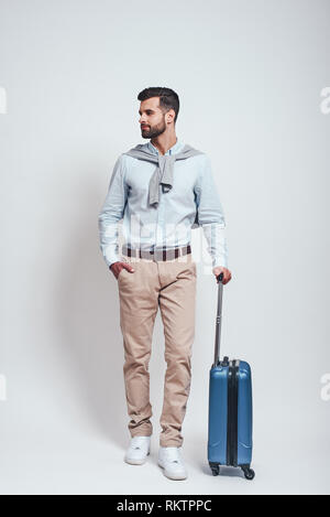 Bereit zu Reisen. Volle Länge von Stattlichen bärtiger junger Mann mit einem Koffer halten eine Hand in der Tasche weg schauen. Grauer Hintergrund. Travel Concept. Stockfoto