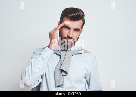 Ups ... bärtiger junger Mann in Freizeitkleidung ist berührend Pflaster auf der Stirn und an der Kamera beim Stehen auf einem grauen Hintergrund. Schmerzen Konzept Stockfoto