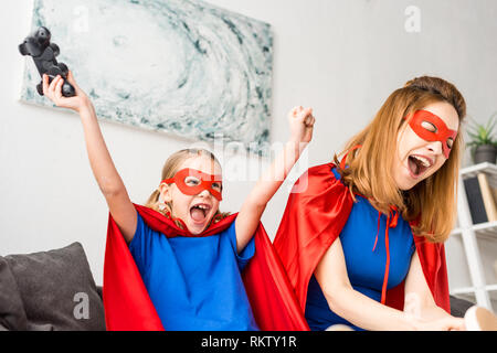 Mutter und Tochter in rot Masken und Umhänge spielen video Spiel zu Hause Stockfoto