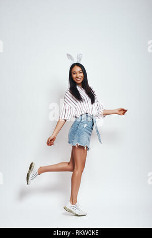 Cute Bunny. Volle Länge des attraktiven lächelnden Asiatin im bunny Ohren- und Casualwear auf grauem Hintergrund. Glück Konzept Stockfoto