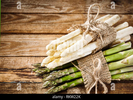 Frische rohe grüne und weiße Spargelspitzen in Bundles mit string und Hessischen zusammen auf rustikalem Holz mit Kopie Raum gestapelt gebunden Stockfoto