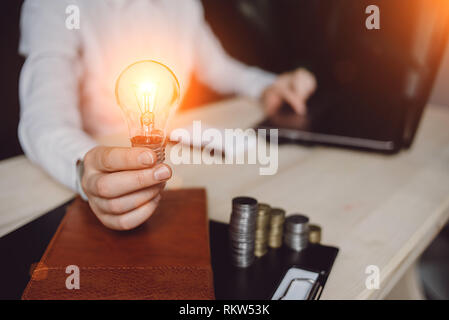 Das Geschäft von Frau Hand, die Lampe mit einem Notebook und Notebook und Geld. Idee, Energieeinsparung und Accounting Finance. Digitale marketin Stockfoto