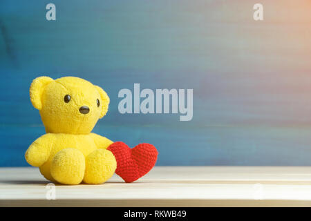Hand Garn machen Rotes Herz neben gelben Teddybär auf Holztisch und blauem Hintergrund. Konzept der Valentines Tag. Stockfoto