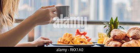 Eine junge Frau mit Frühstück auf dem Balkon. Frühstück mit Kaffee Obst und Brot, Croissant auf einem Balkon vor dem Hintergrund der großen Stockfoto