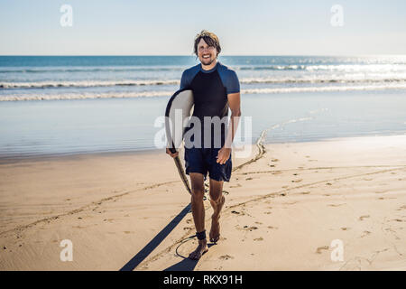 Schöne sportliche junge Surfer mit seinem Surfboard unter dem Arm in seinem Anzug posiert an einem Sandstrand der tropischen Strand Stockfoto