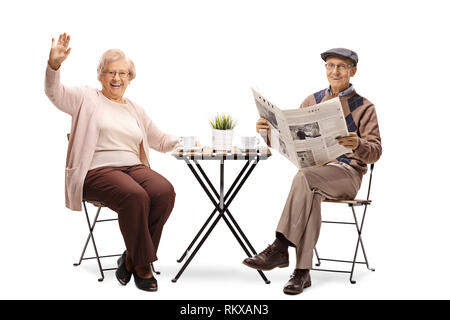 In voller Länge Porträt einer älteren Frau an einem Tisch sitzen mit einer Tasse Kaffee und winken und ein älterer Mann eine Zeitung lesen isoliert auf weißem b Stockfoto