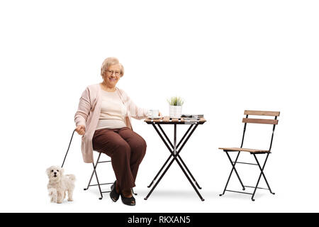 In voller Länge Porträt einer älteren Frau an einem Tisch sitzen mit einer Tasse Kaffee und ihrem Hund neben dem Stuhl auf weißem Hintergrund Stockfoto