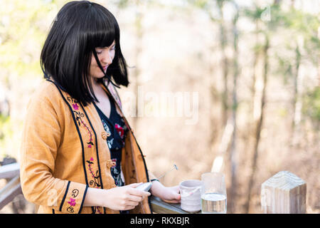 Glas Wasser und einer Tasse Tee auf hölzernen Geländer auf Hinterhof deck Garten und fröhliche Frau mit Schneebesen für Matcha, schwarze Haare Mädchen und asiatischen Tuch Stockfoto