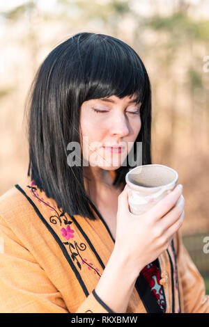 Frau Mädchen schwarze Haare holding Matcha Grüntee cup schweren closeup Trinken nach aussen im Garten im Hinterhof mit geschlossenen Augen Stockfoto