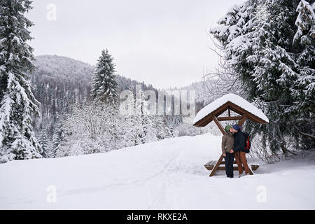 Paar in Wanderausrüstung küssen beim Stehen durch eine Zuflucht in einer verschneiten Clearing während heraus für eine Winterwanderung gemeinsam Zärtlich Stockfoto