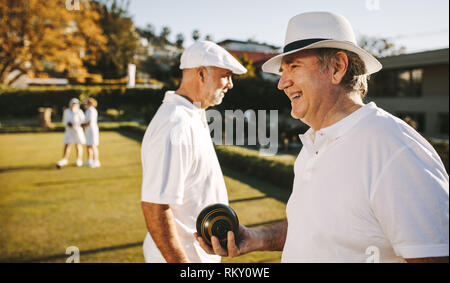 Seitliche Sicht auf einen alten Mann in einem Park Boccia an einem sonnigen Tag. Älterer Mann in Hut Boccia in eine Wiese mit Freunden. Stockfoto